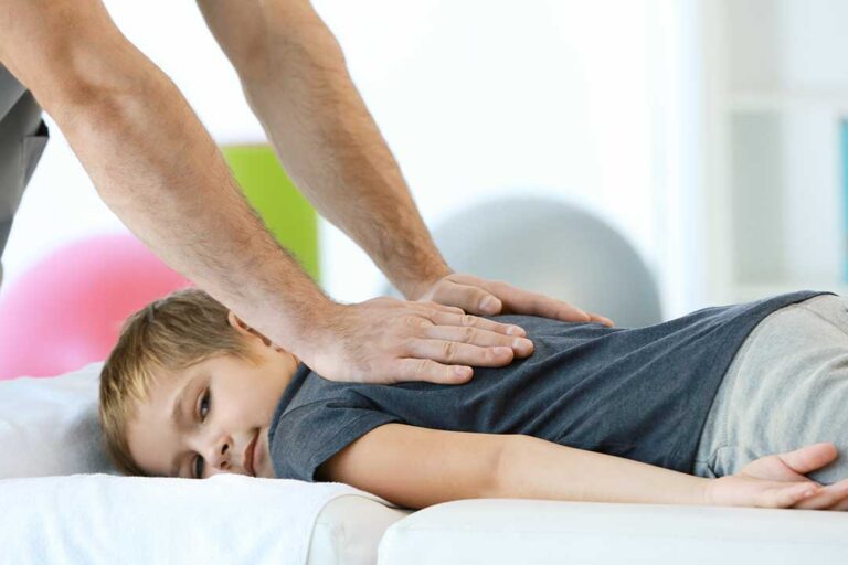 A pregnant woman in comfortable clothing sits on a padded chiropractic table talking to a healthcare provider.